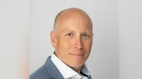 Supplied A head and shoulders image of Mark Edwards. He is wearing a light grey jacket over a white shirt. He is smiling and looking into the camera.