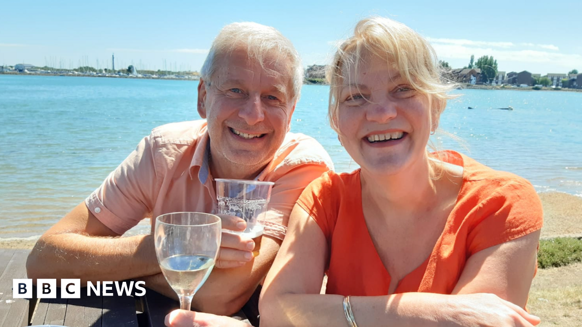 Chris and Ruth Stone-Houghton sit on a picnic bench, smiling into the camera. They are holding alcoholic drinks, and behind them is a body of water