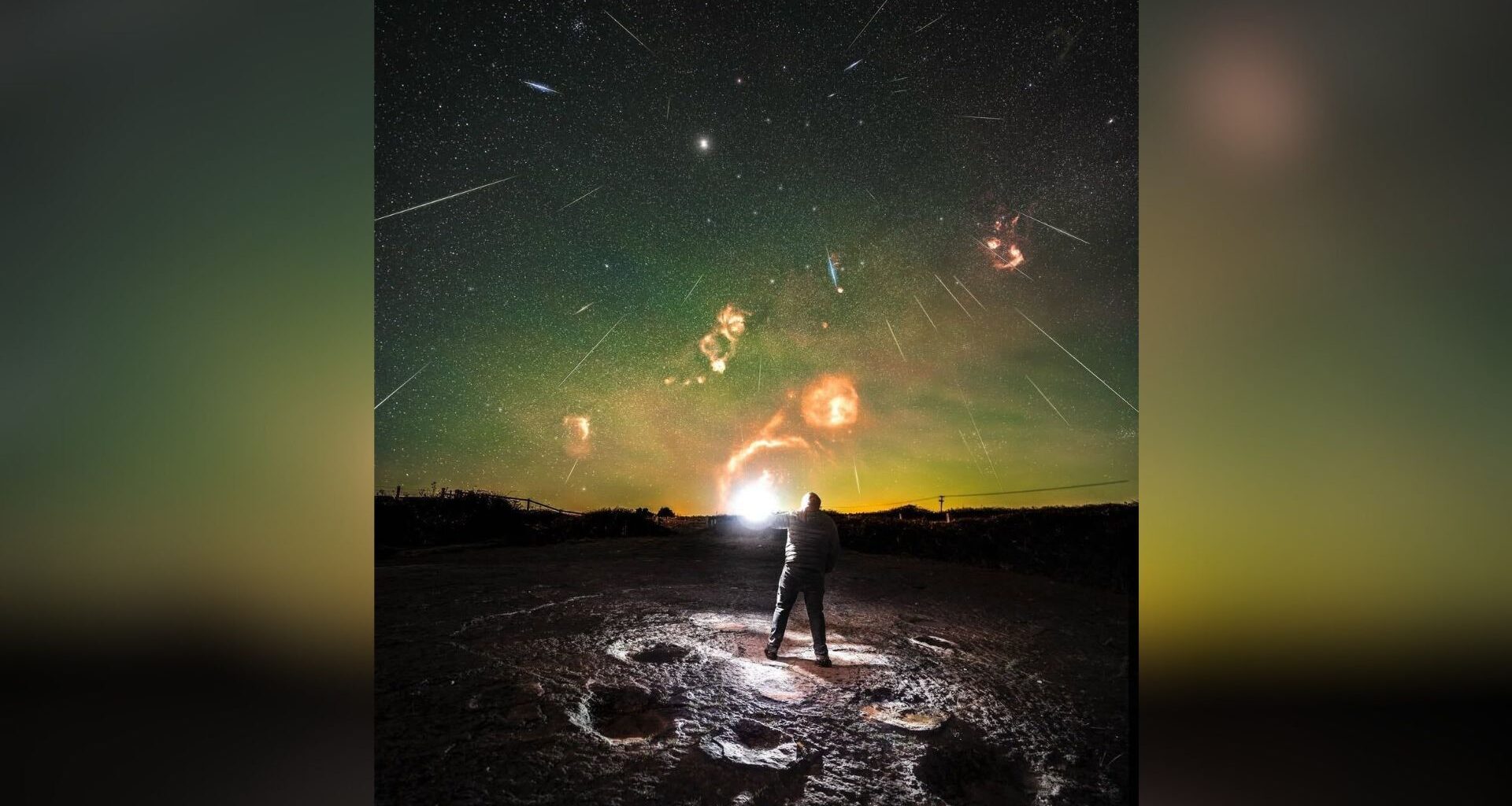 A person standing on a rock looks up at a green night sky with streaks of light showing a meteor shower overhead