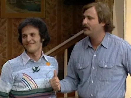Two people in a living room. One man wears a patterned pastel sweater; the other has a mustache and wears a button-up shirt, gesturing with his thumb