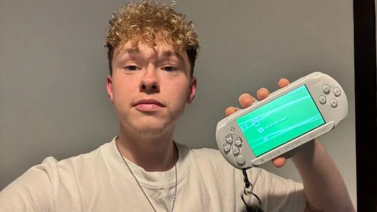Kyle is indoors holding up a white PlayStation Portable (PSP) console with a keychain attached.