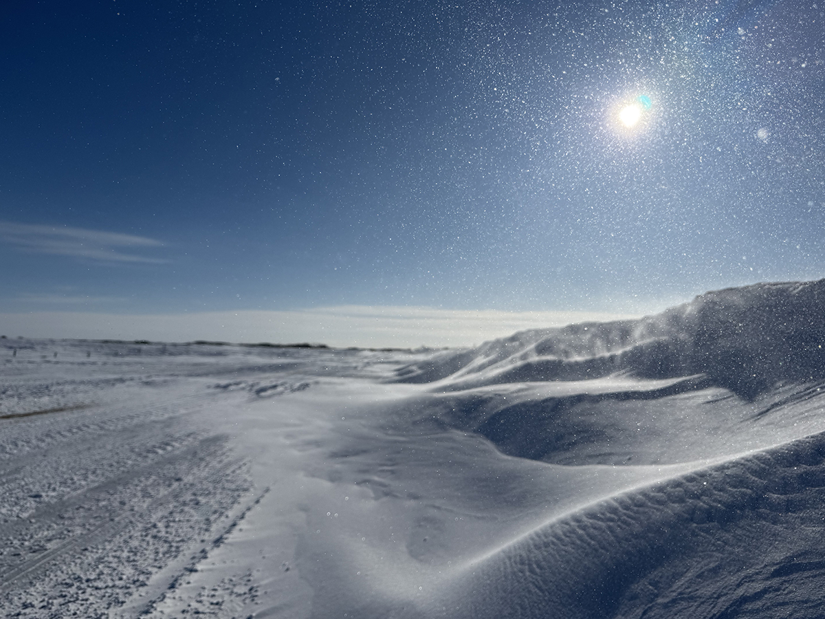 Snow is piled along the side of a road and the sun is low in the clear blue sky on a cold day in February in Saskatchewan.