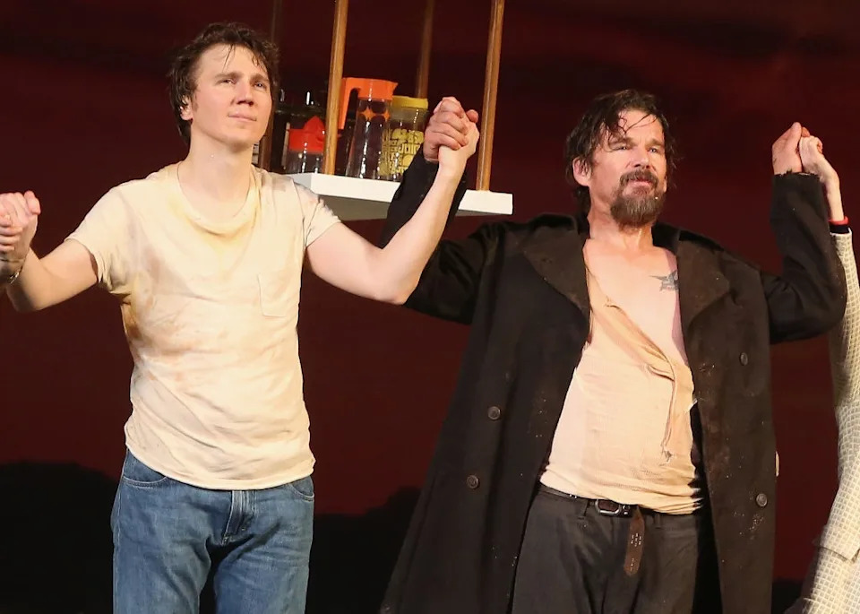Two actors stand on stage holding hands, one in a casual t-shirt and jeans, the other in a long coat with an open shirt, expressing triumph