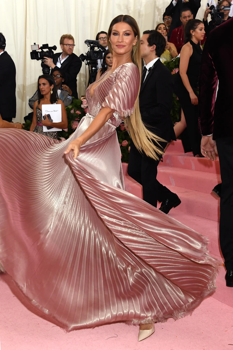 Person twirls in a flowing, pleated gown with structured shoulders, posing on the red carpet at a celebrity event