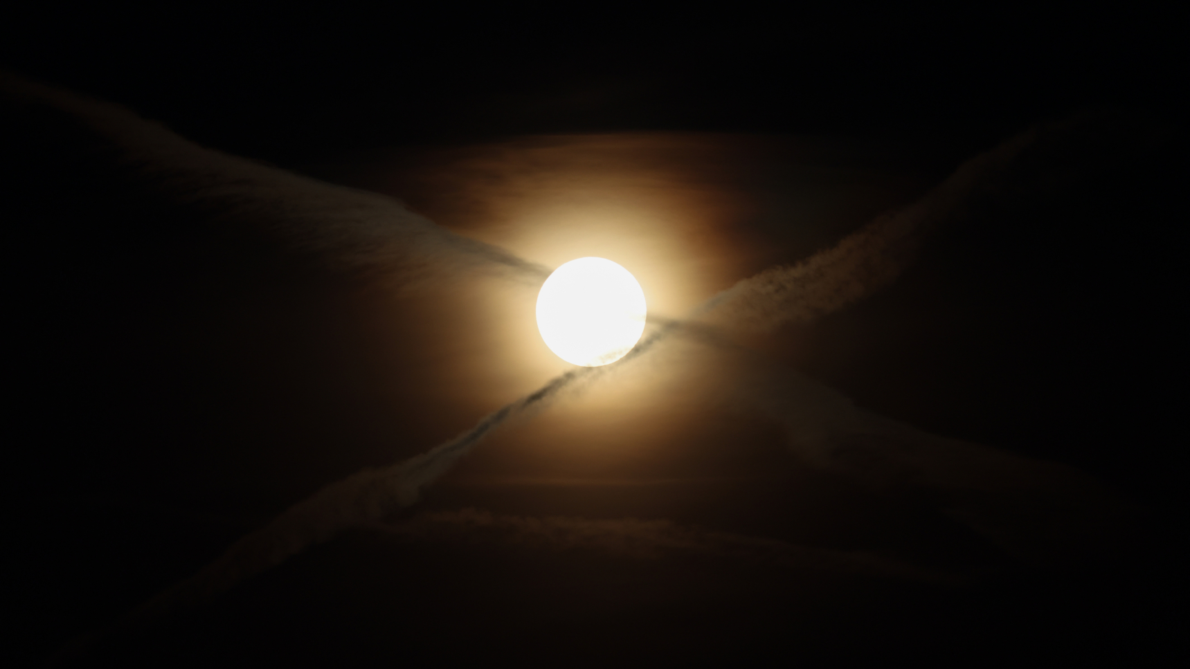 The glowing disk of a full moon is pictured glowing against a black night sky behind a thin veil of cloud as two airplane trails create an 'X' -shape in the sky.
