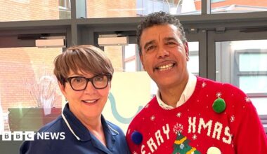 Woman in a blue nurse's outfit, standing on a stone balcony with a glass railing behind. In the background is a stone viaduct and she is surrounded by various shrubs and trees including bright red begonias.