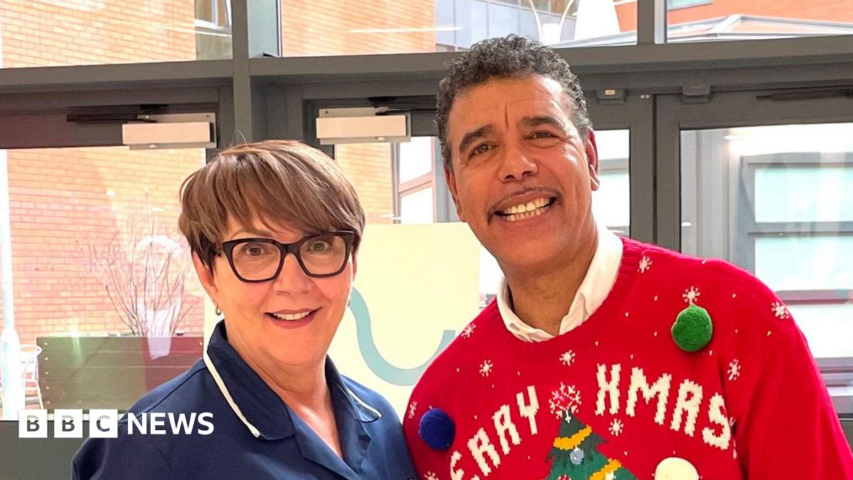 Woman in a blue nurse's outfit, standing on a stone balcony with a glass railing behind. In the background is a stone viaduct and she is surrounded by various shrubs and trees including bright red begonias.