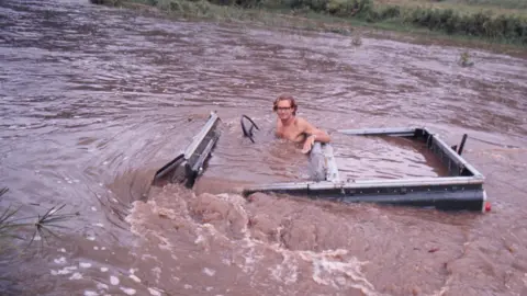 Oria Douglas-Hamilton An old photo of Iain Douglas-Hamilton sitting in an open-topped jeep style vehicle which is almost totally submerged in a brown river. 