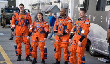 Four astronauts - three men and one woman - wearing orange spacesuits stand on the street. Behind them, people are standing or walking by. There is a partially visible van behind the crew; the van has a picture of the Moon and the words "Artemis II" on it.