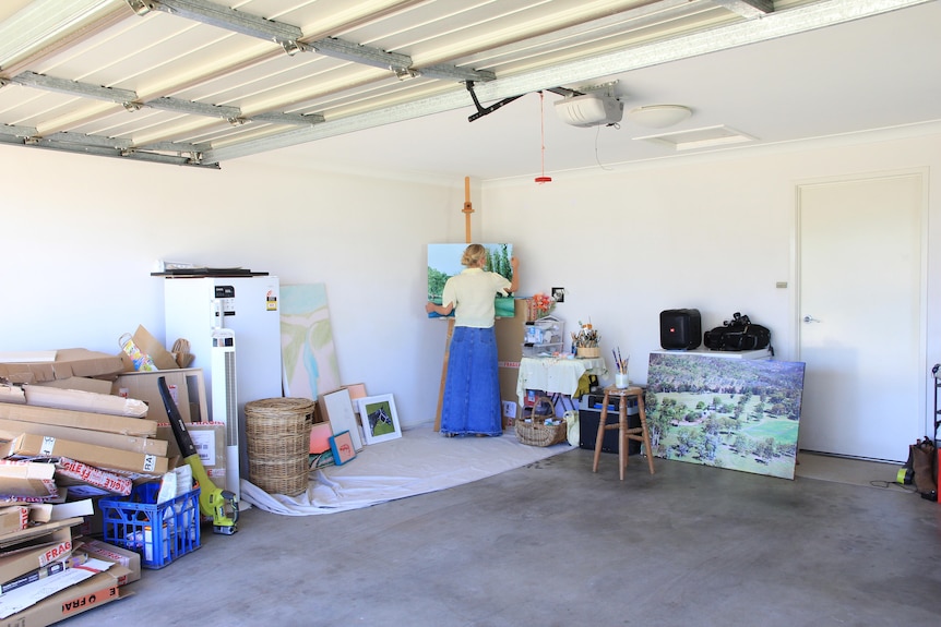 A woman is painting on a canvas in the corner of a garage with lots of debris around her. 