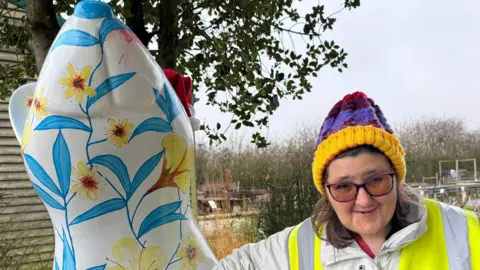 Amber Macey/BBC A woman wearing a brightly-coloured bobble hat, glassed and hi-vis vest standing next to the flowery rat sculpture.