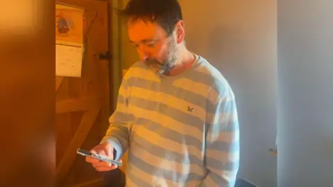 Steven Medway A man with spiky brown hair wearing a blue and white striped jumper and looking down at a movile phone in his hand.