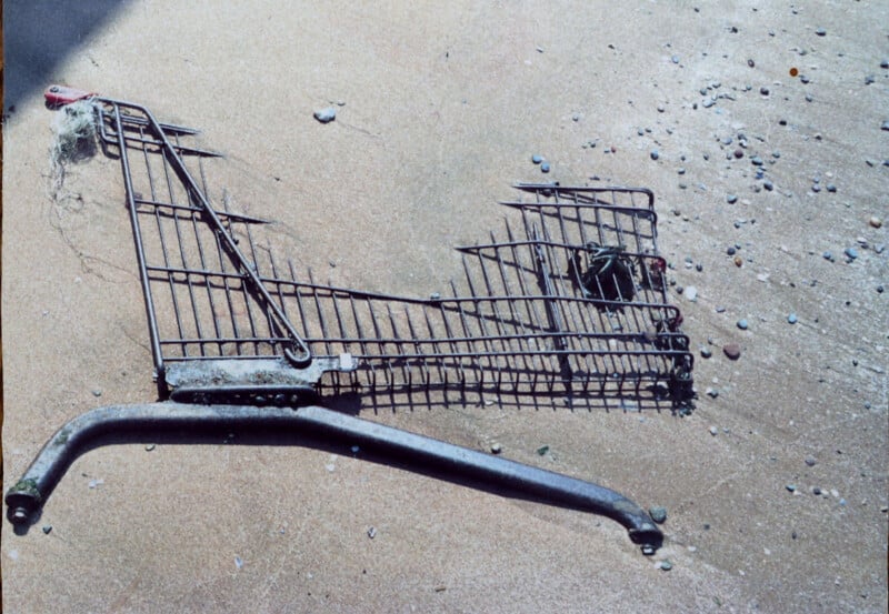 A rusty, broken shopping cart lies partially buried in wet sand on a beach, surrounded by small pebbles and debris.