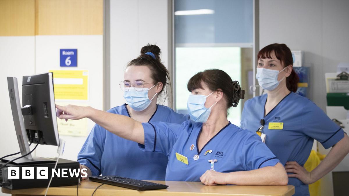 Three nurses work inside a hospital wearing face masks, pointing at a computer monitor.