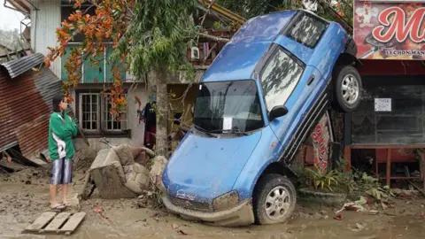 Getty Images A young woman in a green and white jacket looks on forlornly as a blue car hangs from a tree, with its rear end in the air, having been lifted by floods during Typhoon Rai in the Philippines in 2021