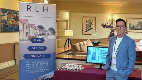 RLH Architectural Nick Cox standing in front of a stall showcasing his company - RLH Architectural.  He is wearing a blue suit jacket and trousers and a white t-shirt.  He has dark hair and glasses.