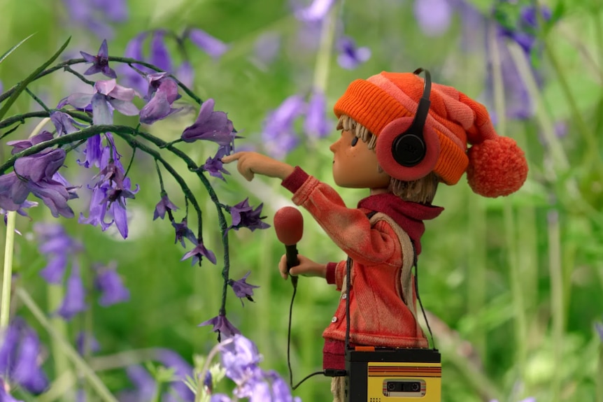 A child holds a microphone and curiously touches a bluebell flower. They're wearing headphones, an orange beanie and hoodie.