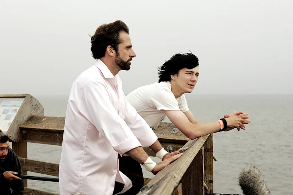 Two people stand on a pier, looking thoughtfully at the ocean. One wears a light dress shirt, and the other a casual tee