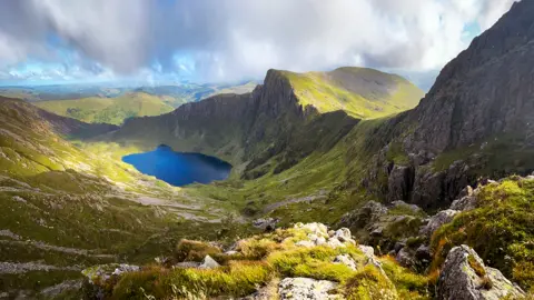 Getty Images Photo taken from near the top of Cader Idris. The angle looks down onto a mountain lake which is bordered on three sides by mountainsides forming a cauldron. The day is a mixture of bright sun and shade but there are misty-edged heavy and dense white clouds above the mountains which look like they could easily turn to rain.
