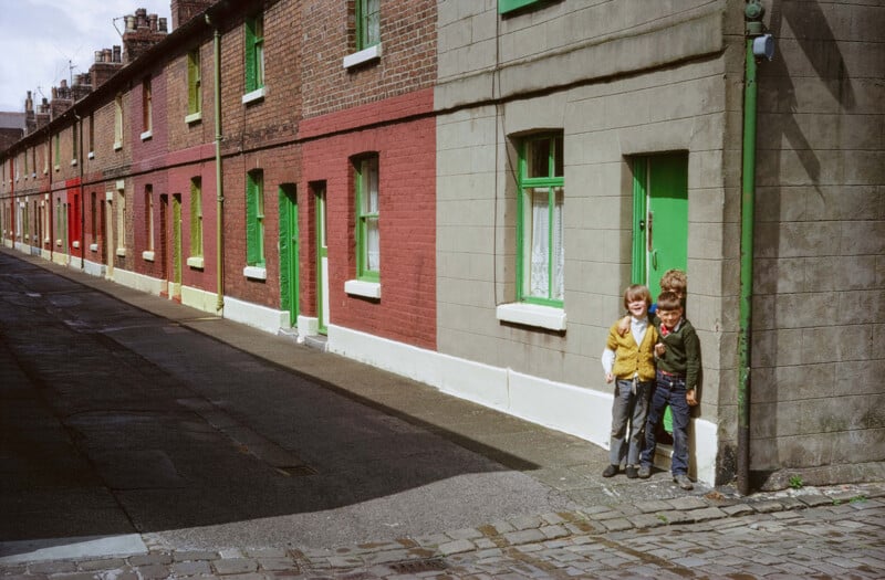 Three children stand together on a quiet cobblestone street outside a row of brick terraced houses with brightly painted green doors and windows. The scene appears to be from a past decade.