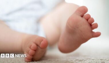 A baby's feet - one on a cream coloured mat or blanket and the other lifted slightly in the air.