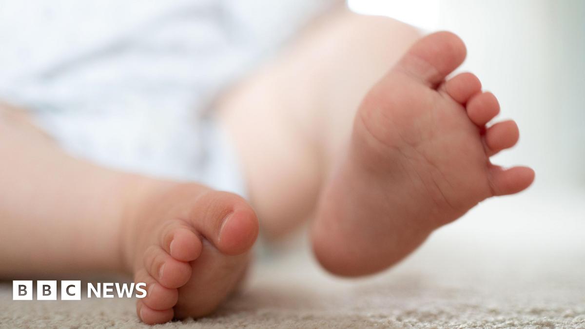 A baby's feet - one on a cream coloured mat or blanket and the other lifted slightly in the air.
