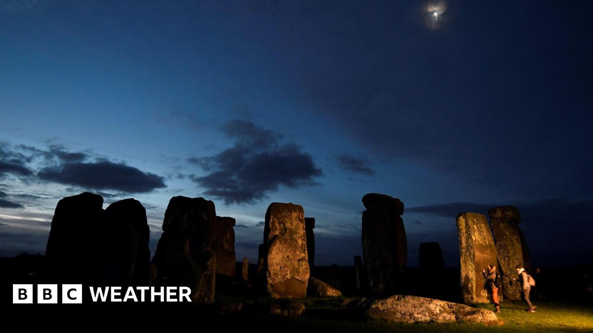 Stonehenge at dawn, with a mainly dark sky with glimmers of brightness and the moon overhead and two people next to the standing stones.