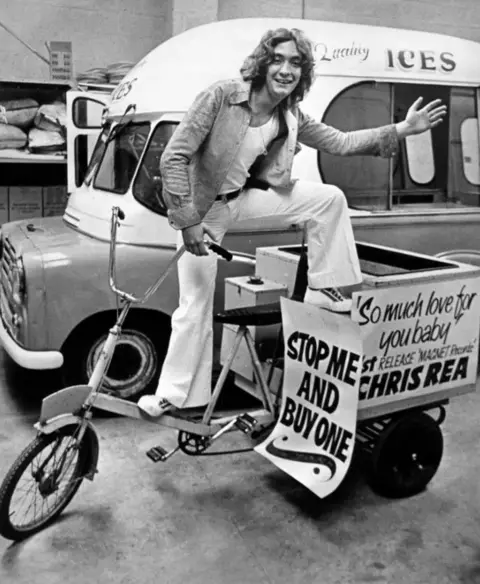 Getty Images Black and white picture of a young Chris Rea posing on a trike and in front of a van selling ice creams. He has long shoulder length hair and wears white flares.