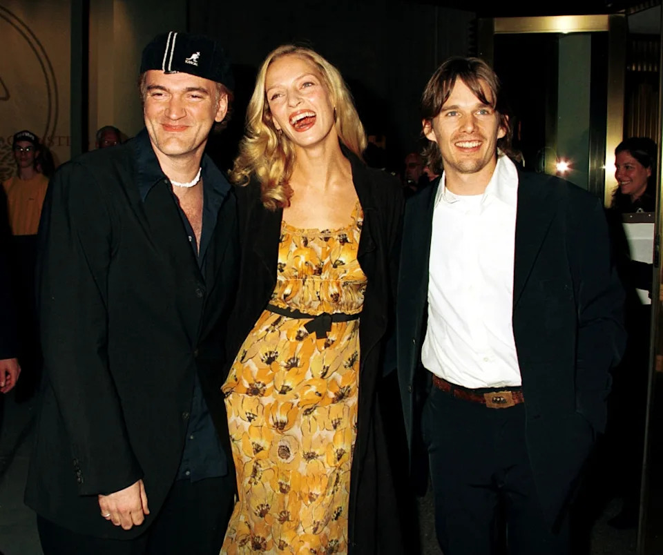 Three individuals at an event; the woman wears a floral dress, flanked by two men in dark suits and casual attire
