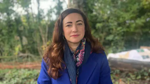 BBC Margaret Gibney sitting outdoors, looking at the camera.  She has long, dark, wavy hair and is wearing a long royal blue coat with a navy and pink scarf and hooped earrings.  There are trees in the background. 