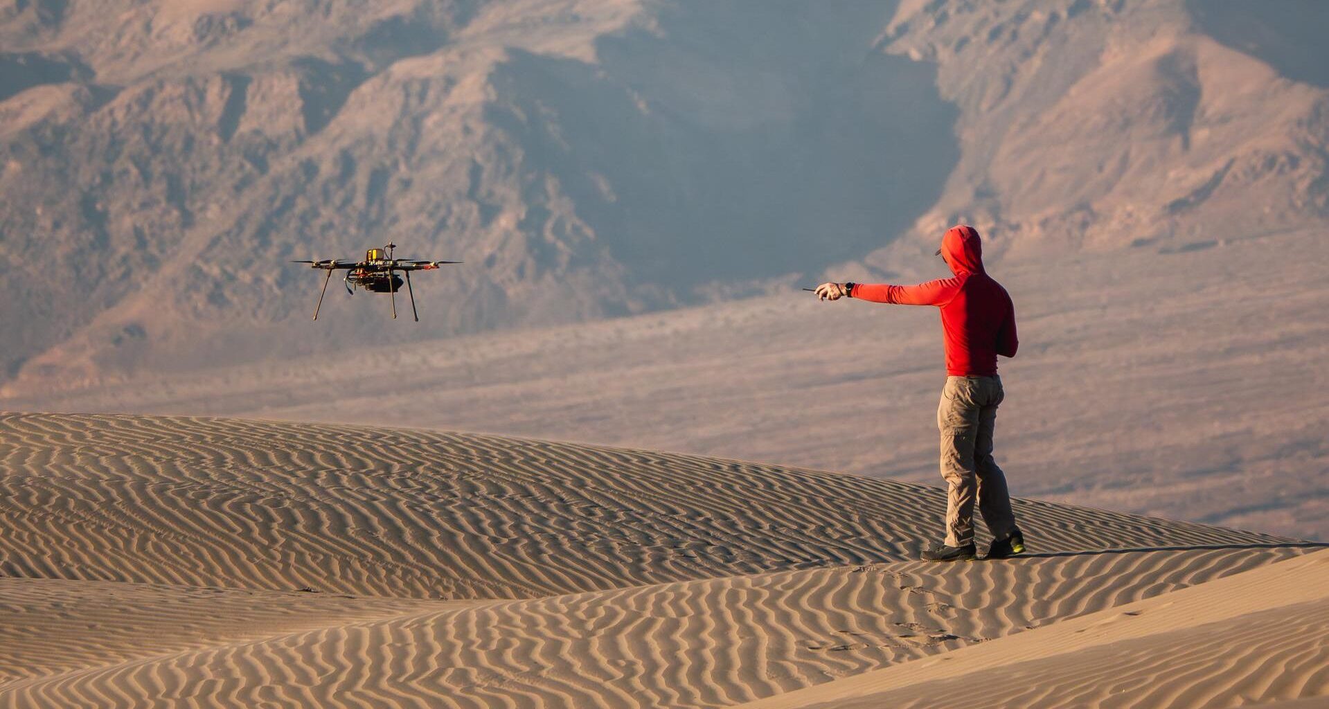 A man wearing a red hoodie and tan pants points to a nearby drone hovering over rippling sand dunes