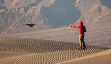 A man wearing a red hoodie and tan pants points to a nearby drone hovering over rippling sand dunes