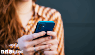 A woman with long red hair and a brown and orange patterned blouse holds a blue smartphone in both hands, as if sending a text message.