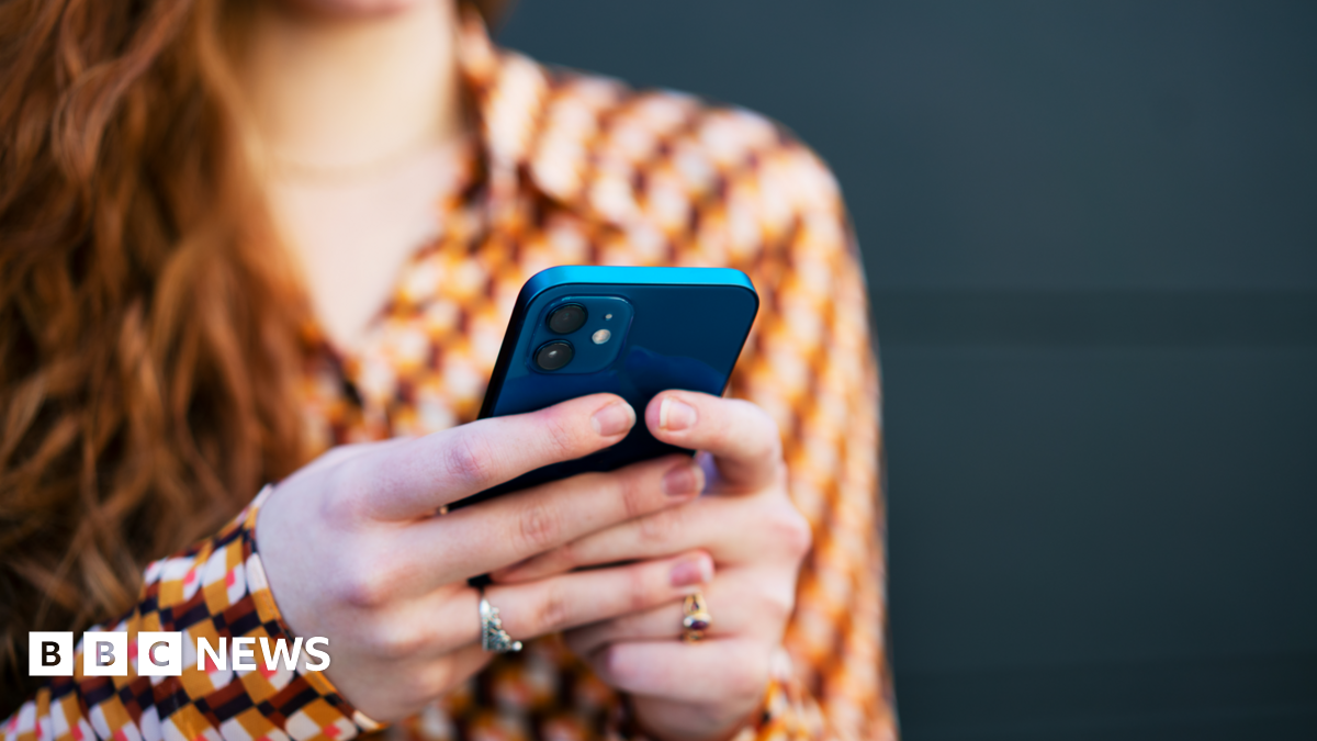A woman with long red hair and a brown and orange patterned blouse holds a blue smartphone in both hands, as if sending a text message.