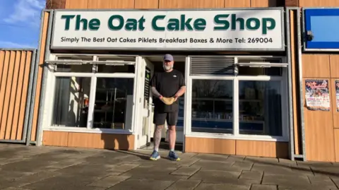 A man in a cap and a black T-shirt and shorts is stood outside a building with a sign across the top saying The Oat Cake Shop