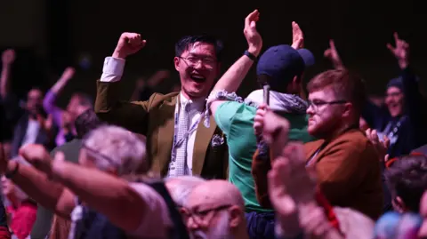 EPA Large crowd of people indoors with raised arms and clenched fists, appearing to celebrate or cheer. Individuals are standing close together, some wearing jackets and scarves, and the background is dimly lit, suggesting a lively event or conference setting.”