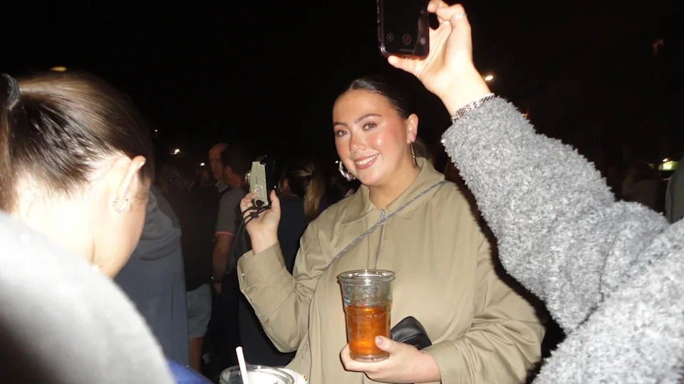 Aoibheann in a beige jacket is at a crowded pub, holding a drink and a small camera, with others around holding phones.