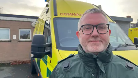 A picture of Neil Sinclair. He is an older man with short, white hair and a white beard. He is smiling at the camera. He is wearing a pair of black, rectangular glasses. Mr Sinclair is standing in front of an ambulance.
