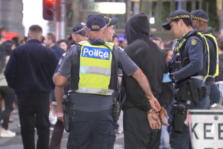 Police officers in navy uniforms with yellow vests that say "police" talk to and walk through crowds of people at night.