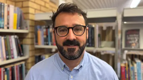 A close-up image of Paul Kelly. He is a young man, with short, dark hair and a dark beard with some patches of grey. He is smiling at the camera. He is wearing a pair of circular black glasses. He is wearing a light blue shirt. He is standing in front of shelves of books, but they are blurred.