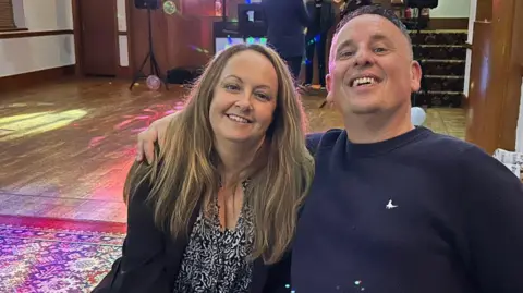 Mark Williams A man and a woman sat, smiling at the camera, with a dance floor, stage and speakers behind them. The woman has long blonde hair and is wearing a black and white blouse and black cardigan. The man, who has short grey hair and is wearing a navy sweatshirt, has his arm around her.