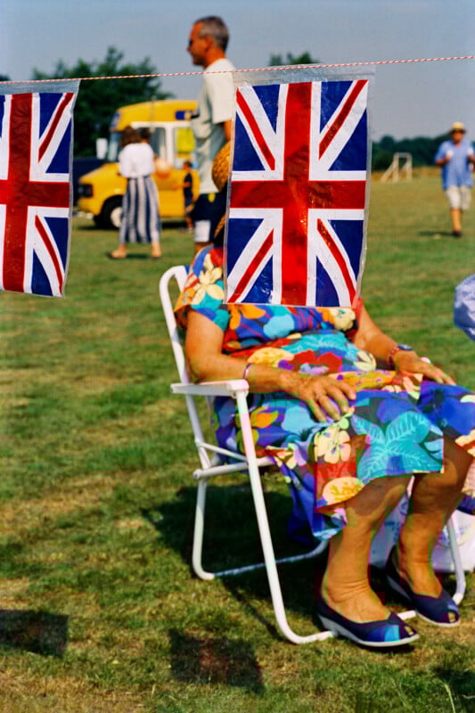 An elderly woman in a colorful dress sits on a lawn chair, her face obscured by a hanging Union Jack flag. People and a yellow van are visible in the background on a sunny day.