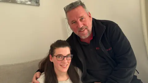 George King/BBC Hannah and her dad Peter sitting on a grey sofa. Hannah is wearing glasses and Peter is wearing a black jacket over a black polo shirt. He is perched on the arm of the sofa and has his arm around Hannah. They are both looking into the camera. 
