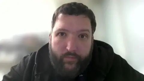 A screenshot of Dr Michael Perry on a online interview. It shows him from the shoulders up. He is wearing a black hoodie, and he has short, dark hair and a dark beard. He is staring at the camera with a neutral expression on his face.