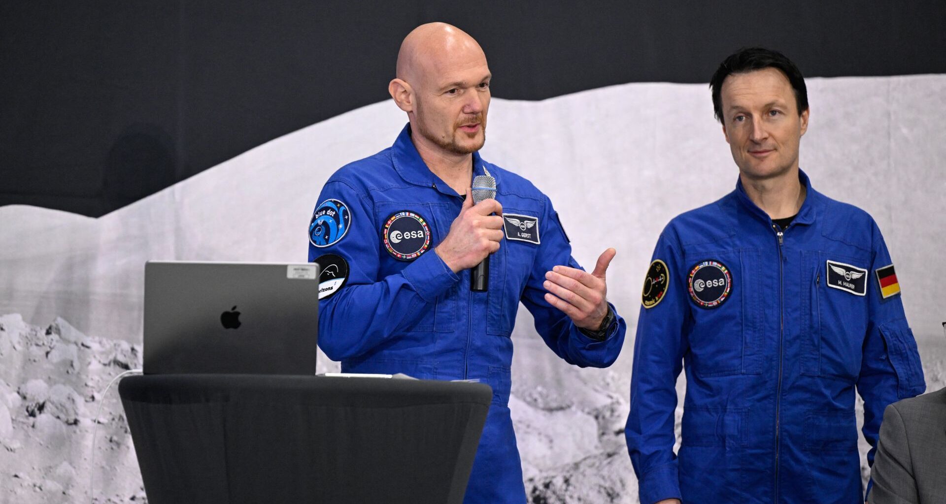 Two astronauts in blue flight suits talk into microphones at a lectern