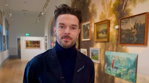 BBC Oliver Jeffers is a man with black hair and beard who is standing in an art gallery in front of a wall of artworks. He is wearing a navy turtleneck top and coat. 