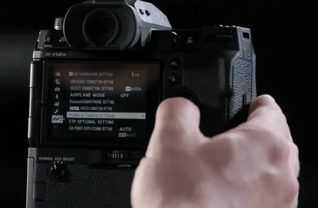 A close-up shot of the back of a black Fujifilm X-H2S camera shows the camera's LCD screen displaying the "NETWORK/USB SETTING" menu, with the option "Frame.io Camera to Cloud" highlighted and selected