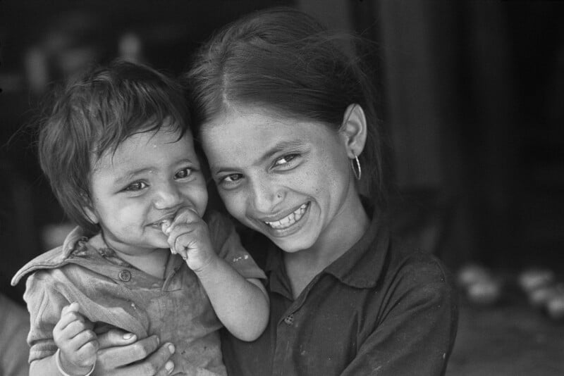 A smiling young girl holds a small child in her arms. Both are looking at the camera; the child is sucking on their fingers while the girl has an arm around them and is wearing hoop earrings. The image is in black and white.