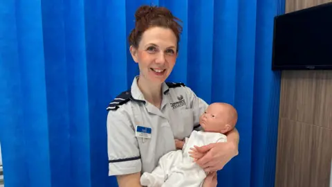Hope Jezzard A woman with brown hair wearing a nurses uniform holding a plastic baby. 