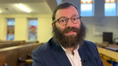 Rabbi Schmuli Brown is sitting in a pew at a synagogue. He has short dark hair, is wearing glasses, and has a long brown beard.